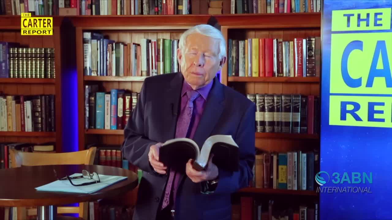 A man in a suit stands in front of a bookshelf, holding a book open. He appears to be reading from the pages.

