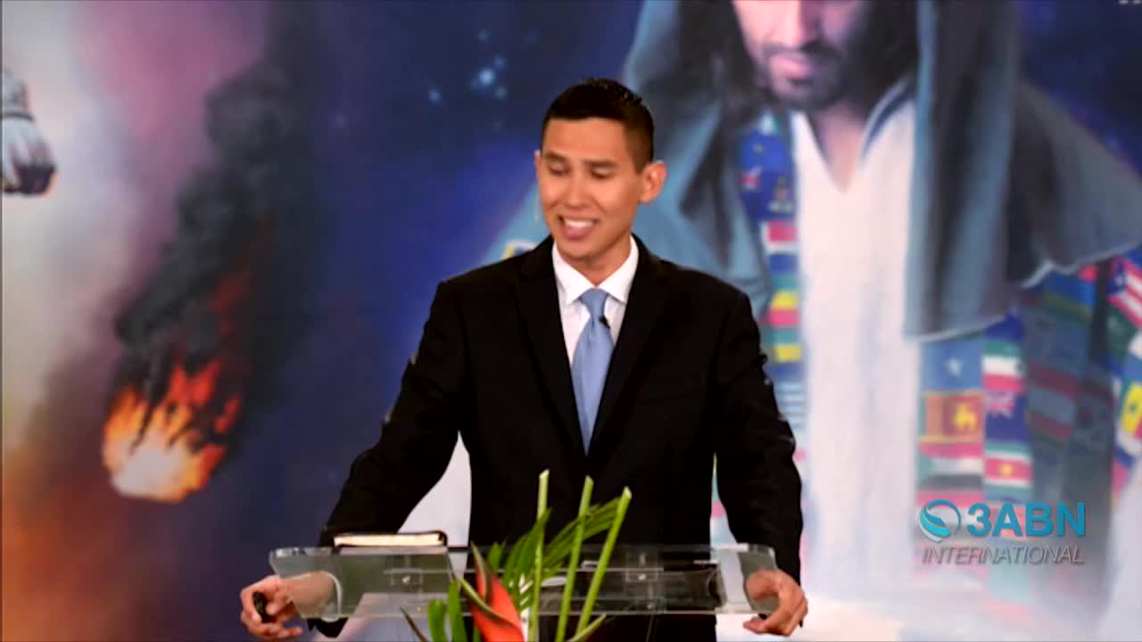 A man in a suit stands at a podium, speaking with a smile. Behind him, a backdrop features a depiction of a figure and a logo for 3ABN International.
