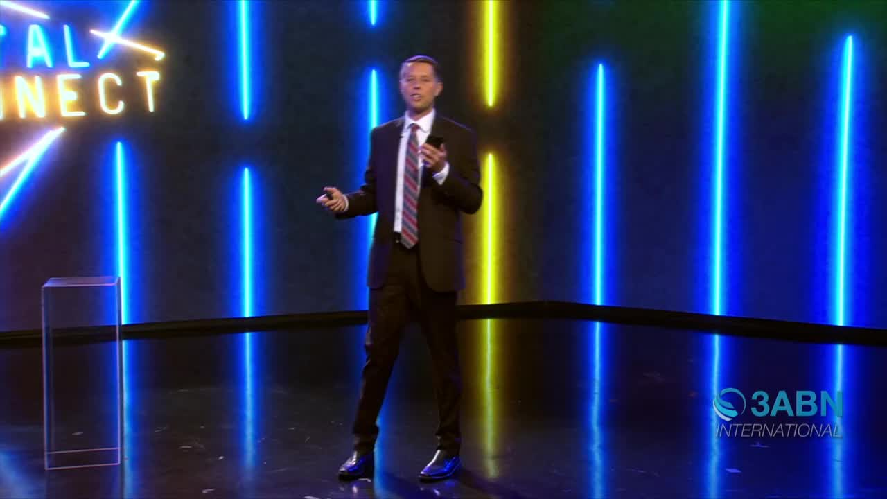 A man in a suit stands on a stage, gesturing with his hands as he speaks. Behind him, vertical blue and yellow neon lights illuminate the dark backdrop, and the 3ABN International logo is visible.
