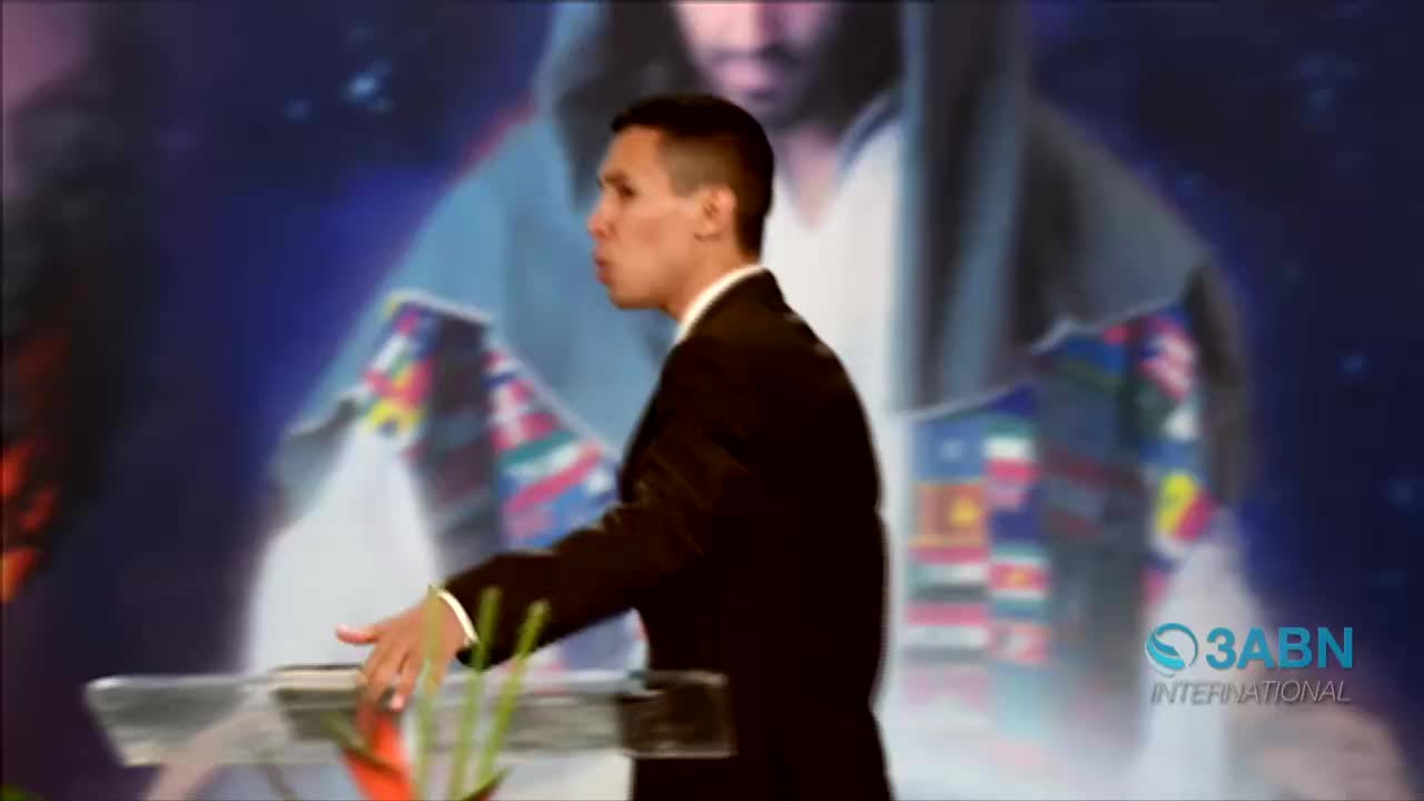A man in a dark suit gestures with his right hand while speaking. Behind him, a blurred image of a figure and flags is visible, along with the 3ABN International logo.
