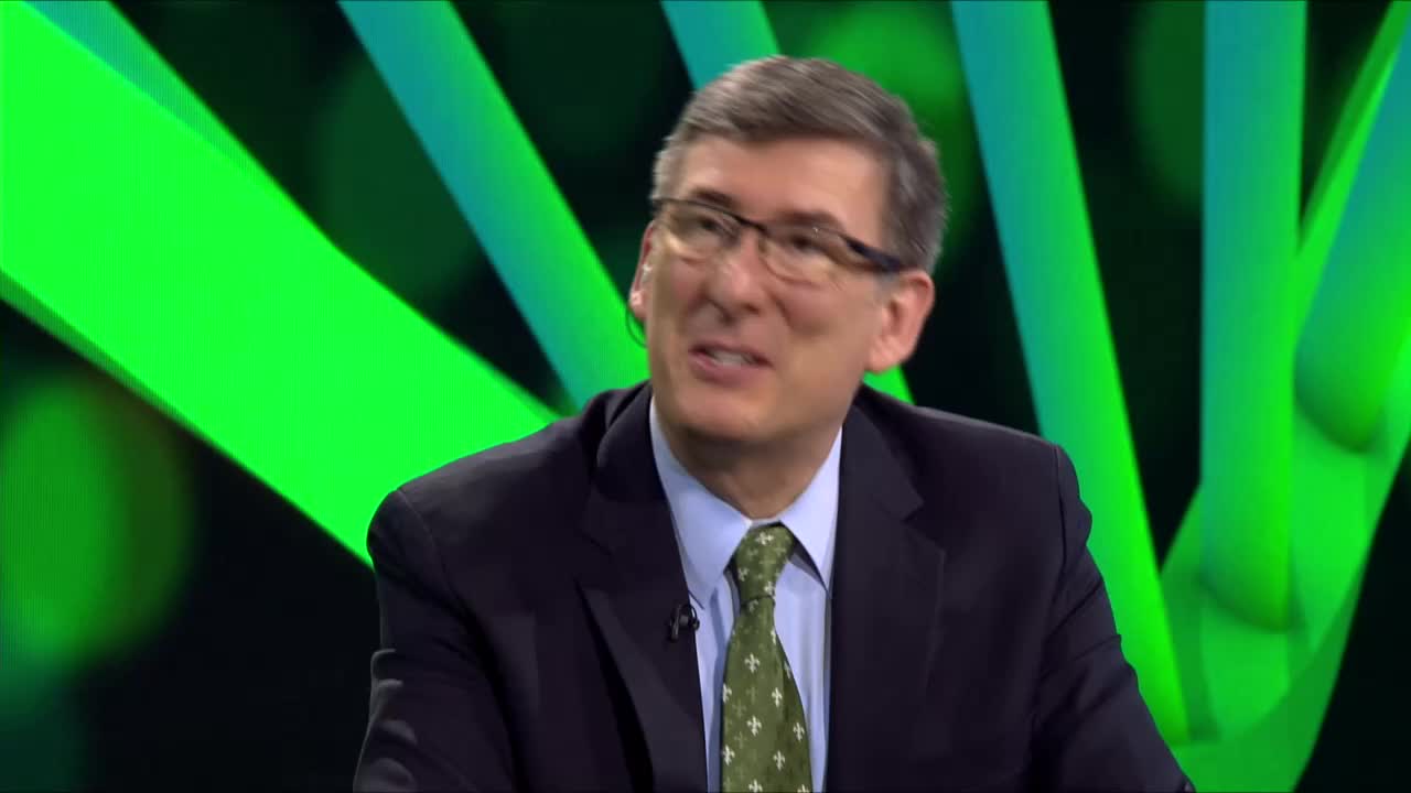 A man in a suit and tie is speaking, his mouth moving as he gestures. Behind him, a bright green backdrop with abstract shapes provides a stark contrast.

