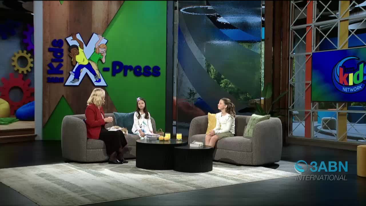 On a set for 3ABN International's Kids X-press, a woman in a red jacket talks to two young girls seated on a gray couch. The girls are listening attentively, with one holding a book in her lap.
