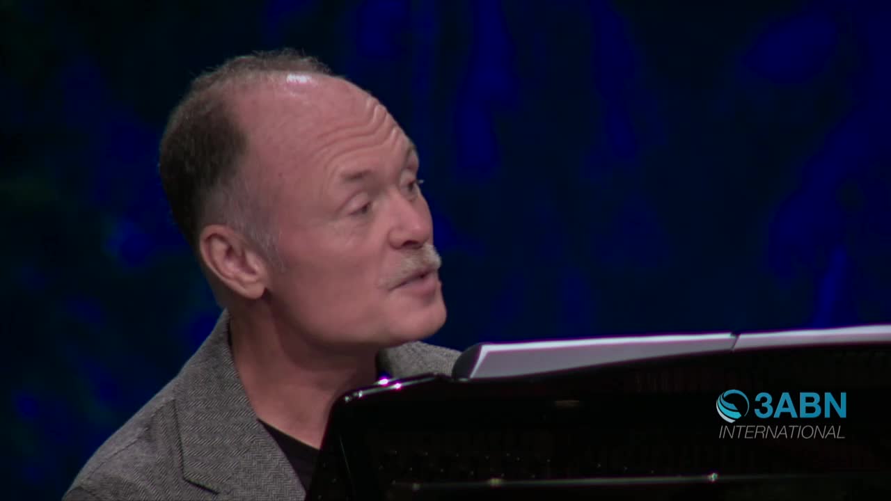 The man with a mustache is speaking, his gaze directed slightly upward. A 3ABN International logo is visible on the piano in front of him.
