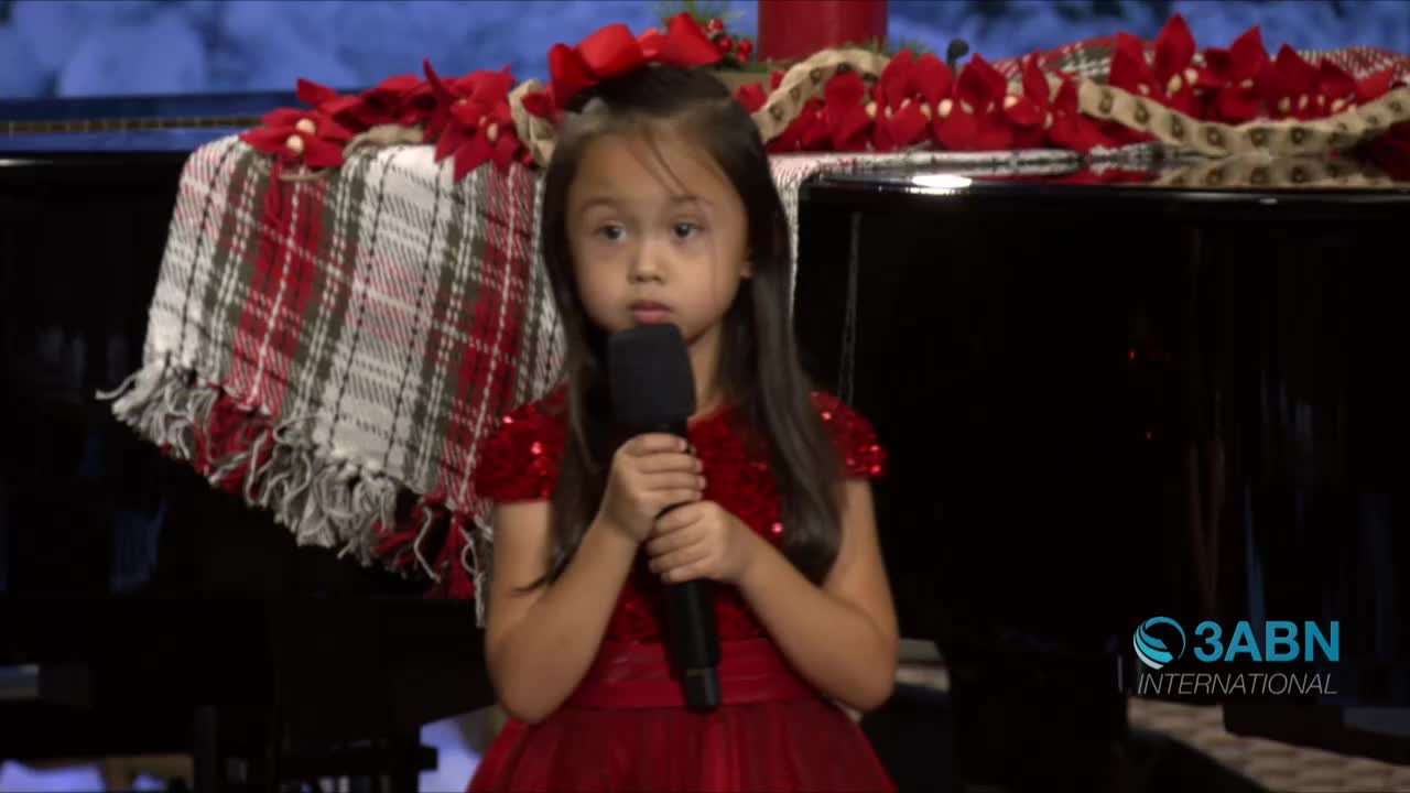 A young girl in a red dress holds a microphone, her gaze fixed forward near a piano decorated for Christmas. The 3ABN International logo is visible on the right.
