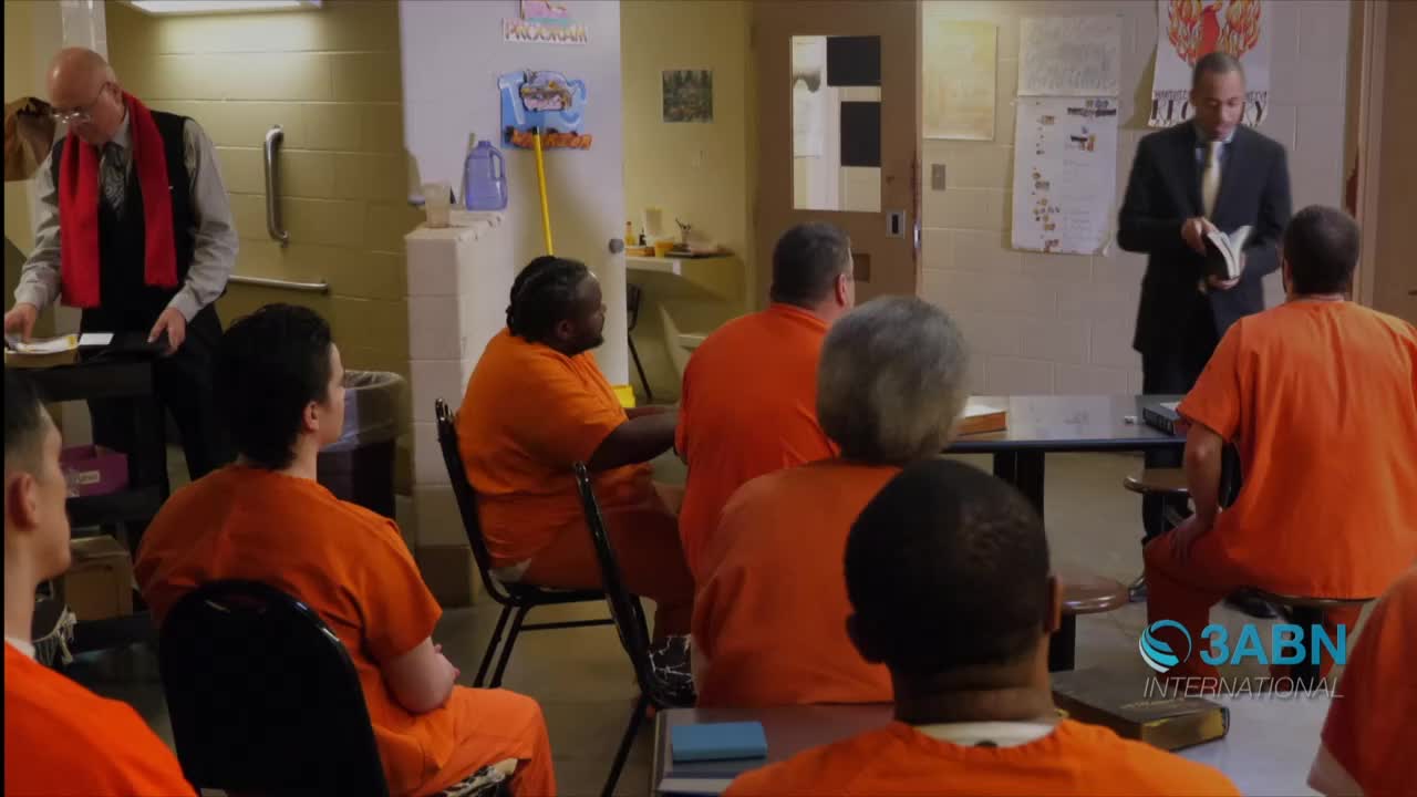 A man in a suit reads from a book to a group of men in orange jumpsuits, seated in a room. Another man, wearing a red scarf, stands near a table, looking at papers.
