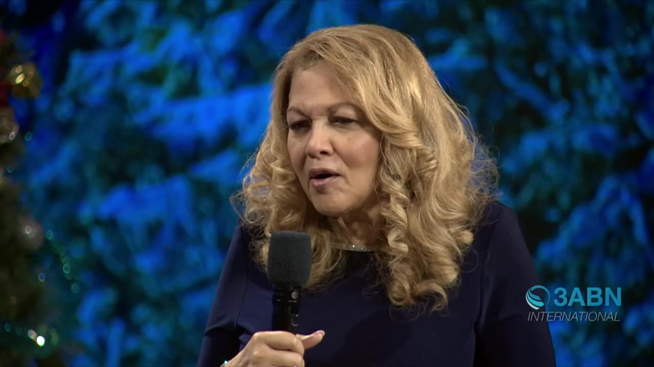 A woman with wavy blonde hair speaks into a microphone, her expression animated. The 3ABN International logo is visible in the corner, and a blue, blurred backdrop is behind her.
