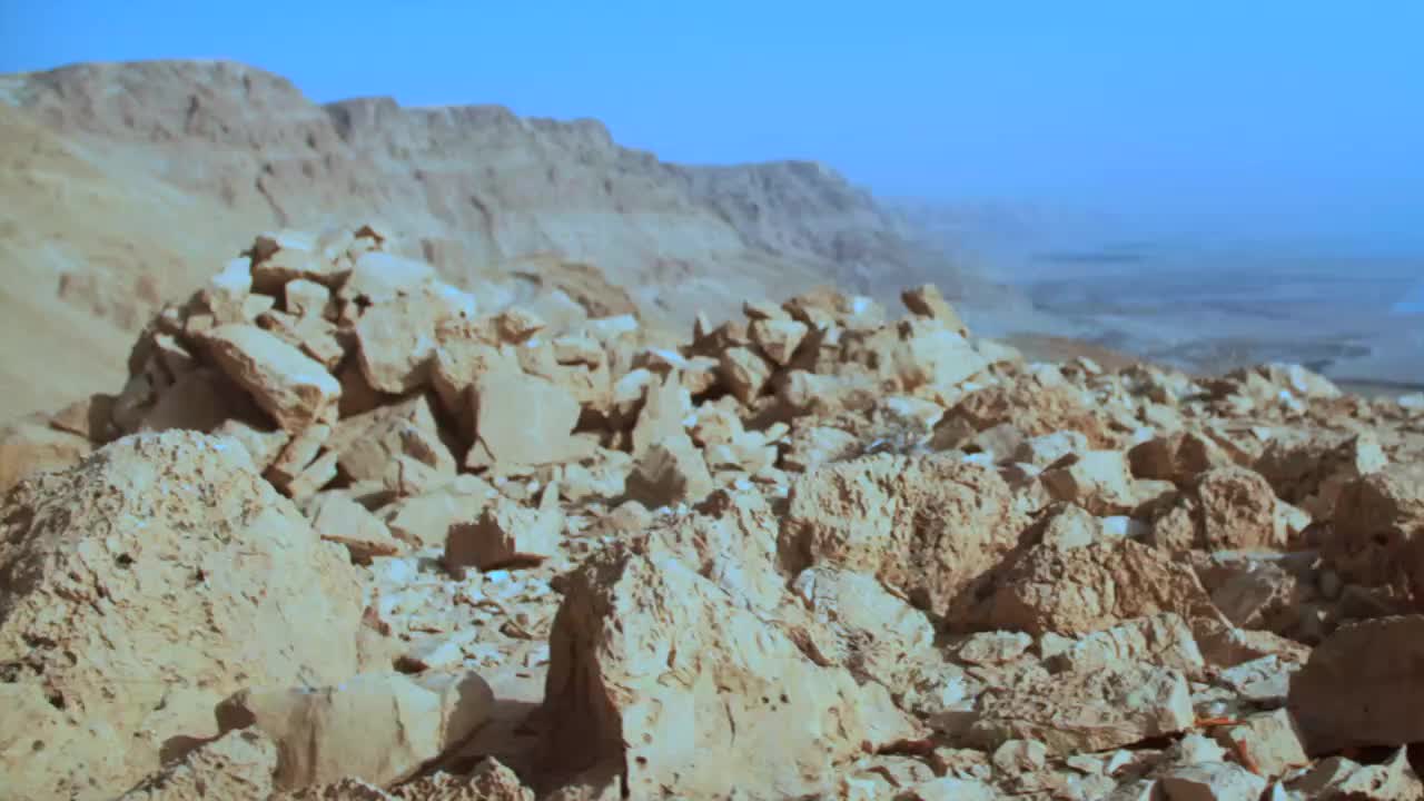 The desert stretches out under a clear blue sky, with rugged, tan mountains rising in the distance. Scattered rocks, some large and jagged, litter the foreground, creating a rough, uneven terrain.