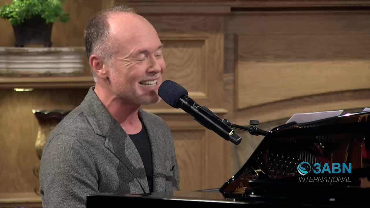 A man with closed eyes and a wide smile sings into a microphone while playing a piano. The 3ABN International logo appears on the piano's polished surface.