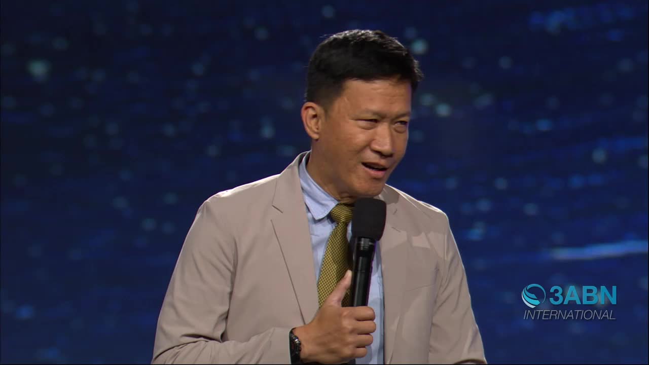 A man in a tan jacket and yellow tie speaks into a microphone on a stage. The 3ABN International logo appears in the lower right corner.