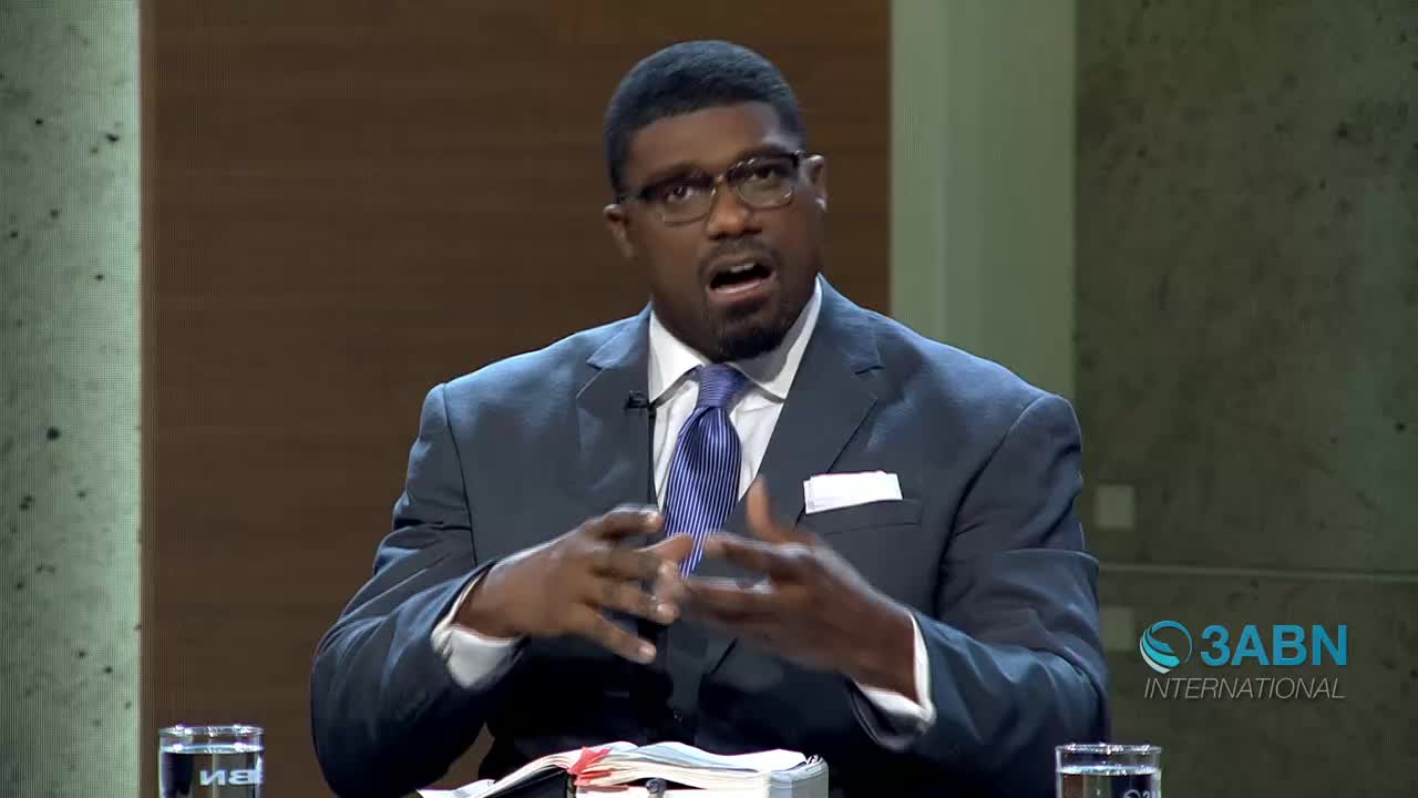 A man in a suit speaks animatedly, gesturing with his hands. A glass of water sits on the table in front of him, beside an open book.