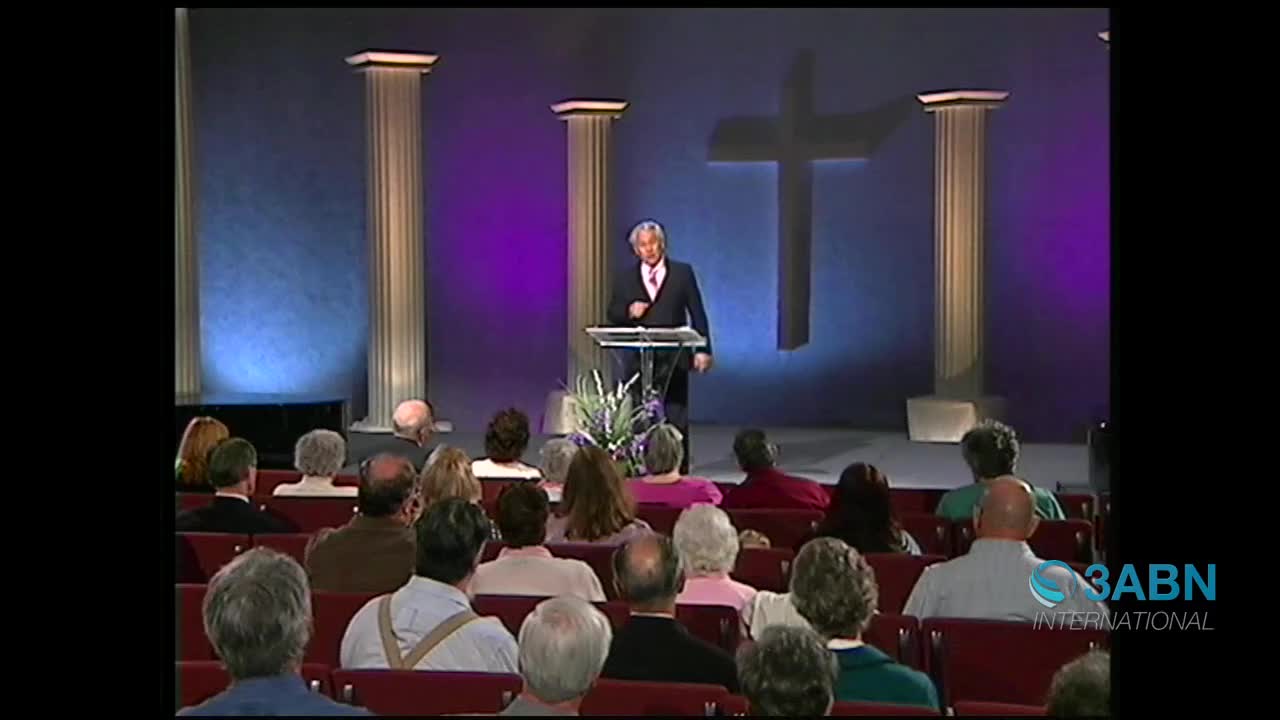 A speaker stands at a podium on a stage, addressing an audience seated in rows. A large cross hangs on the wall behind him, illuminated by soft purple and blue lights.