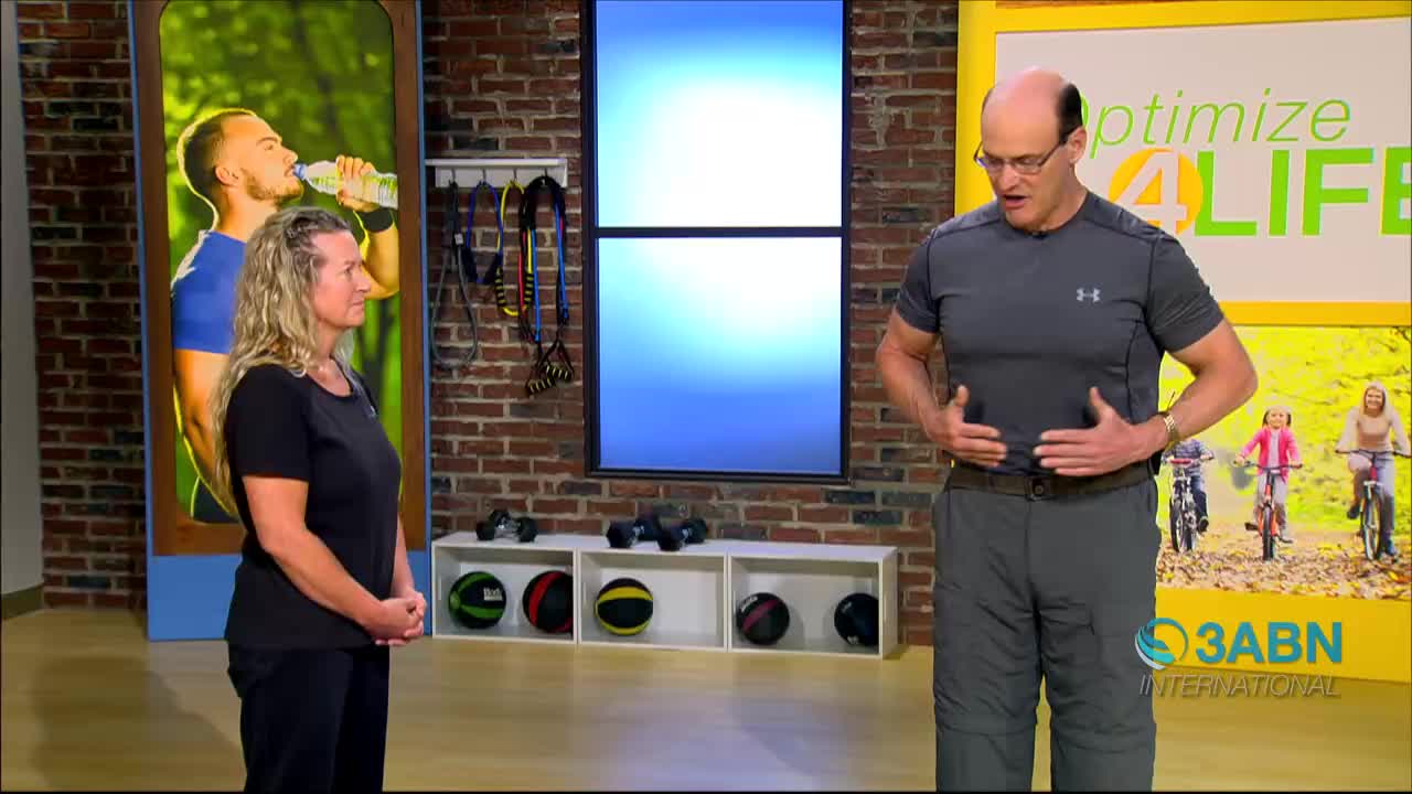 A man in a gray shirt and pants demonstrates an exercise, placing his hands on his abdomen. A woman stands to his left, facing forward. Behind them, a screen displays the 3ABN International logo.