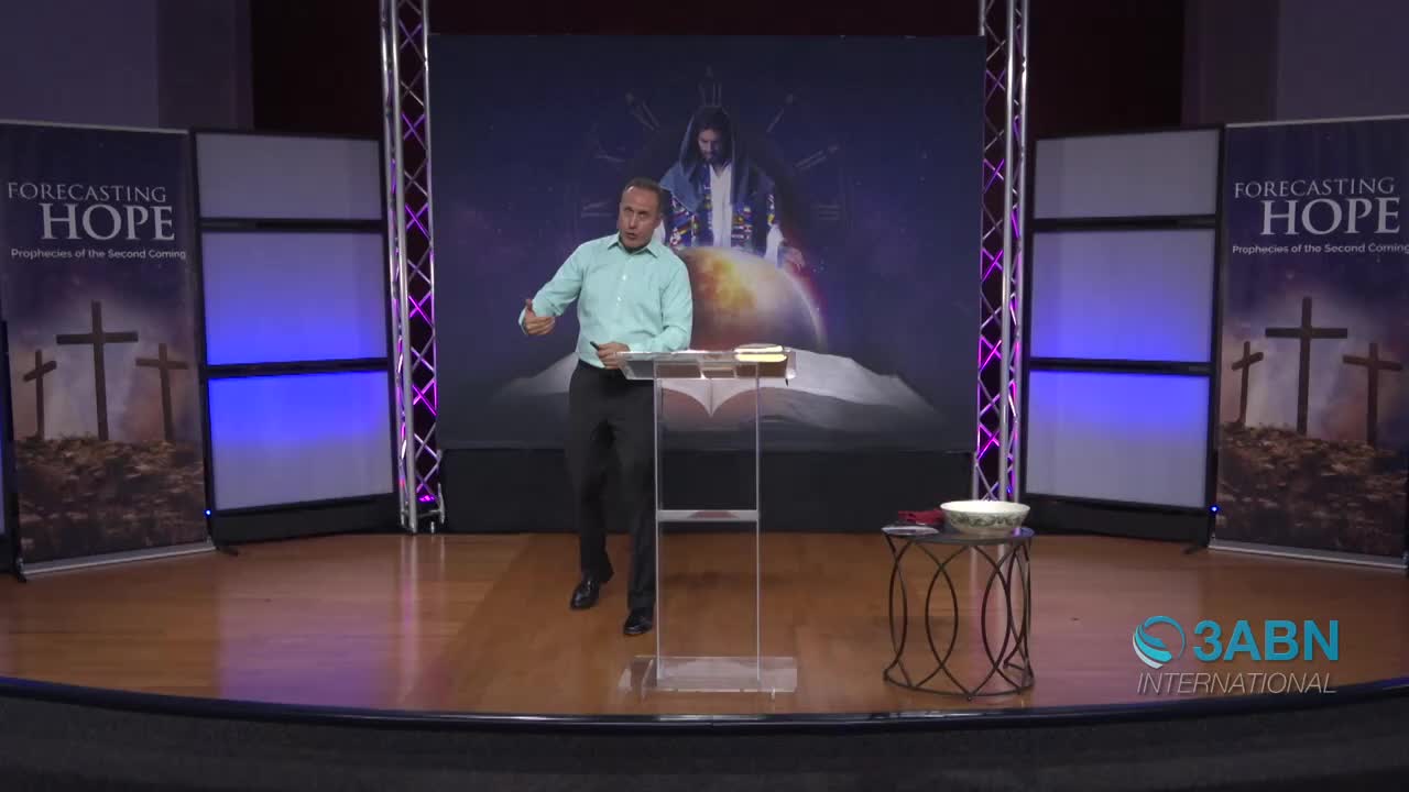 A man in a light blue shirt gestures with his right hand while standing behind a clear podium. The 3ABN International logo appears in the lower right corner of the stage.