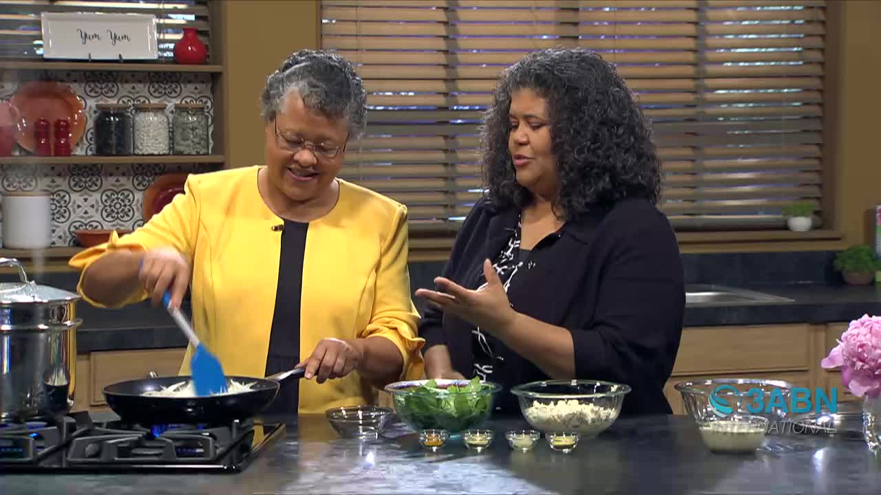 A woman in a bright yellow jacket stirs something in a pan with a blue spatula. Beside her, another woman gestures with her hands, explaining something.
