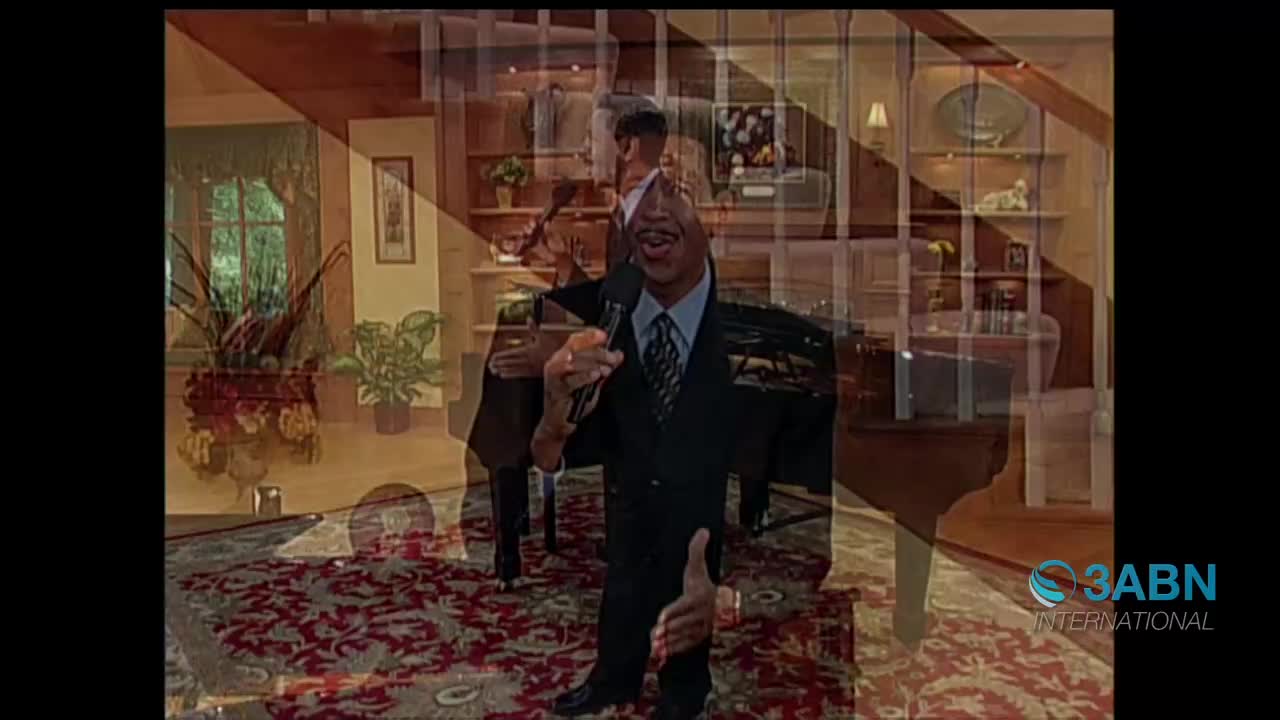 A man in a suit stands holding a microphone, gesturing with his free hand. Behind him, the scene overlays with a grand piano and a staircase.