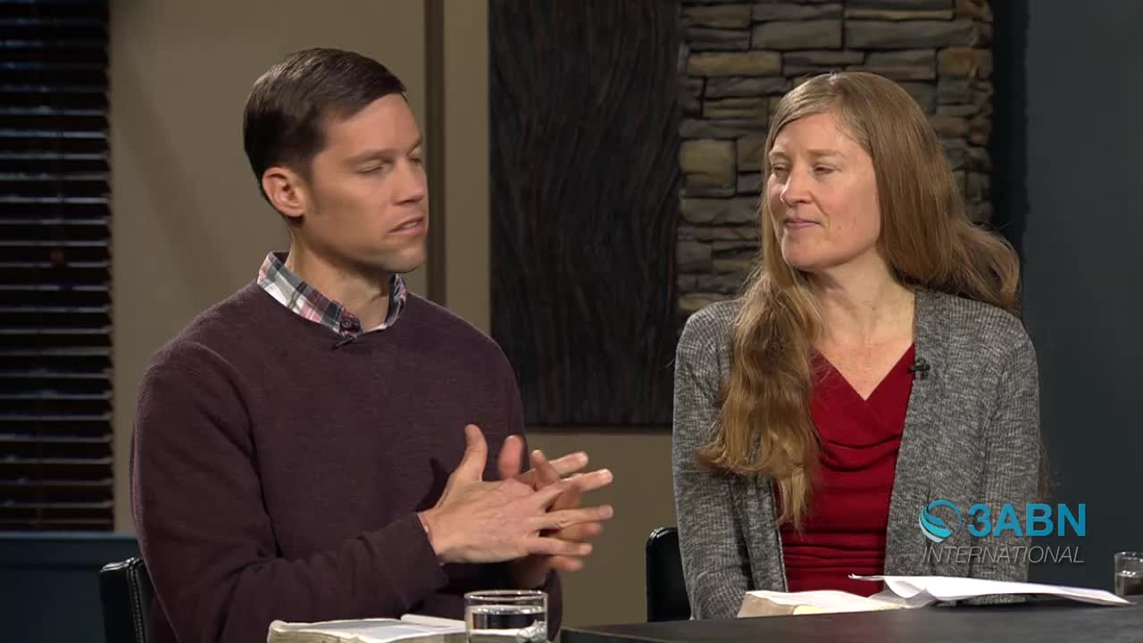 A man with his hands clasped in front of him speaks earnestly. Beside him, a woman with long, light brown hair listens attentively.