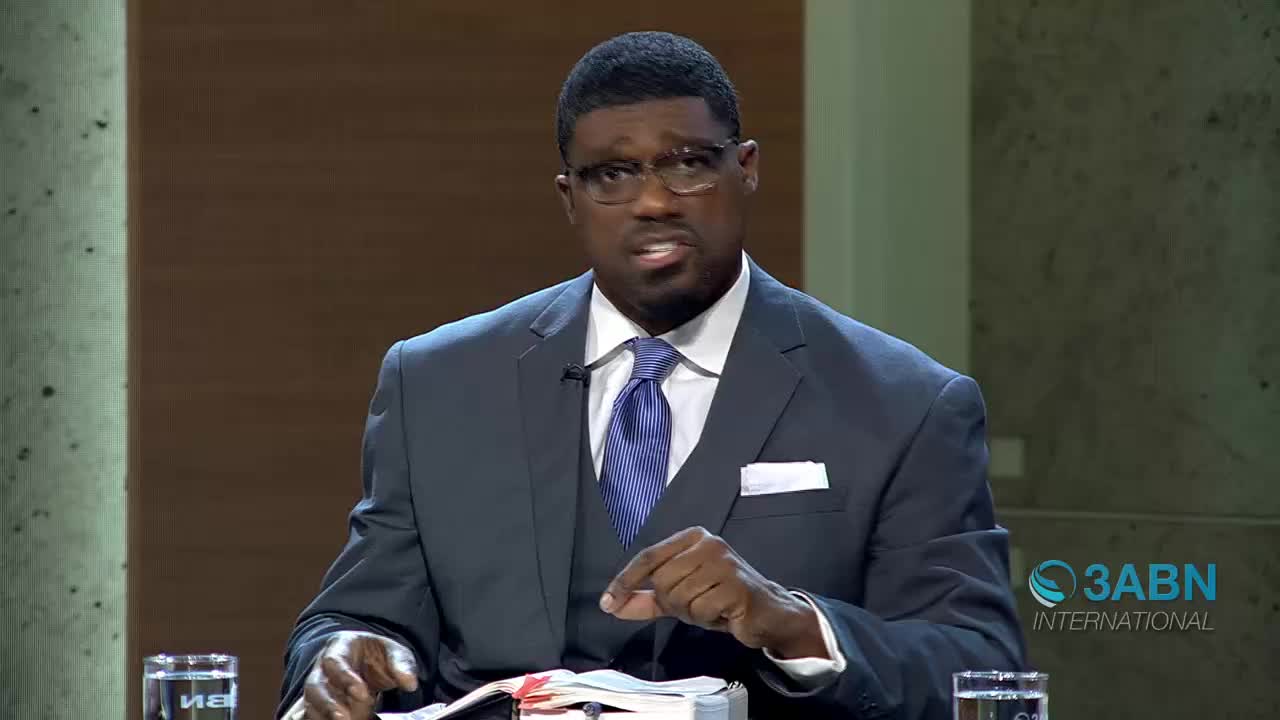 A man in a dark suit and blue tie gestures with his right hand while speaking. He is seated at a table with a Bible in front of him, and the 3ABN International logo is visible in the background.