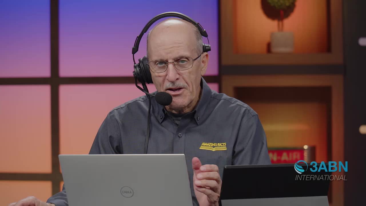 A man with a headset speaks into a microphone, his gaze directed forward. He wears a dark shirt with an "Amazing Facts" logo, and a laptop sits before him.