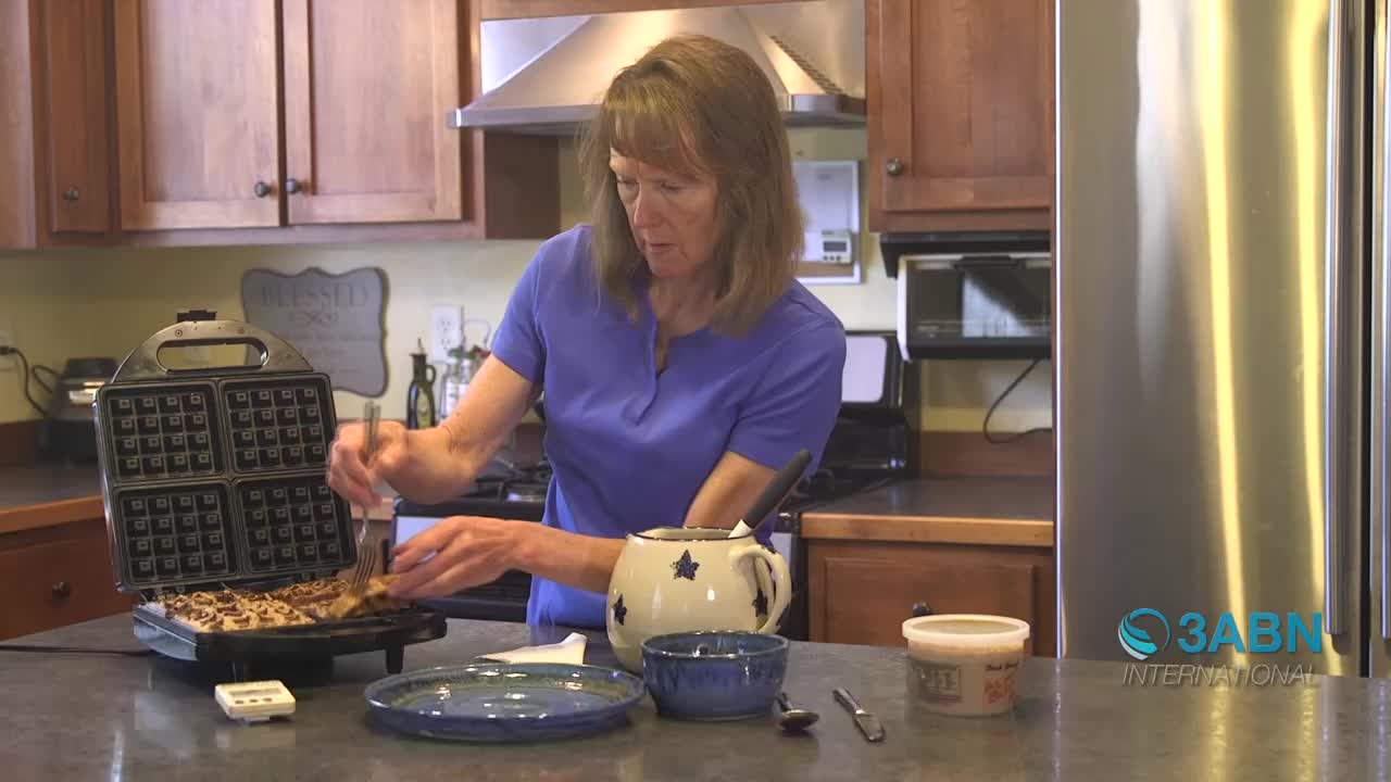 A woman uses a fork to lift a golden-brown waffle from a black appliance. She's in a kitchen, likely preparing breakfast for her family.