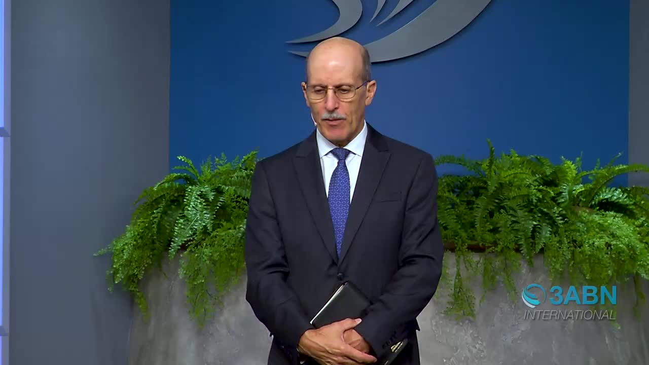 A man in a dark suit stands with his hands clasped around a black book. Lush green ferns frame the stage behind him, where the 3ABN International logo is visible.