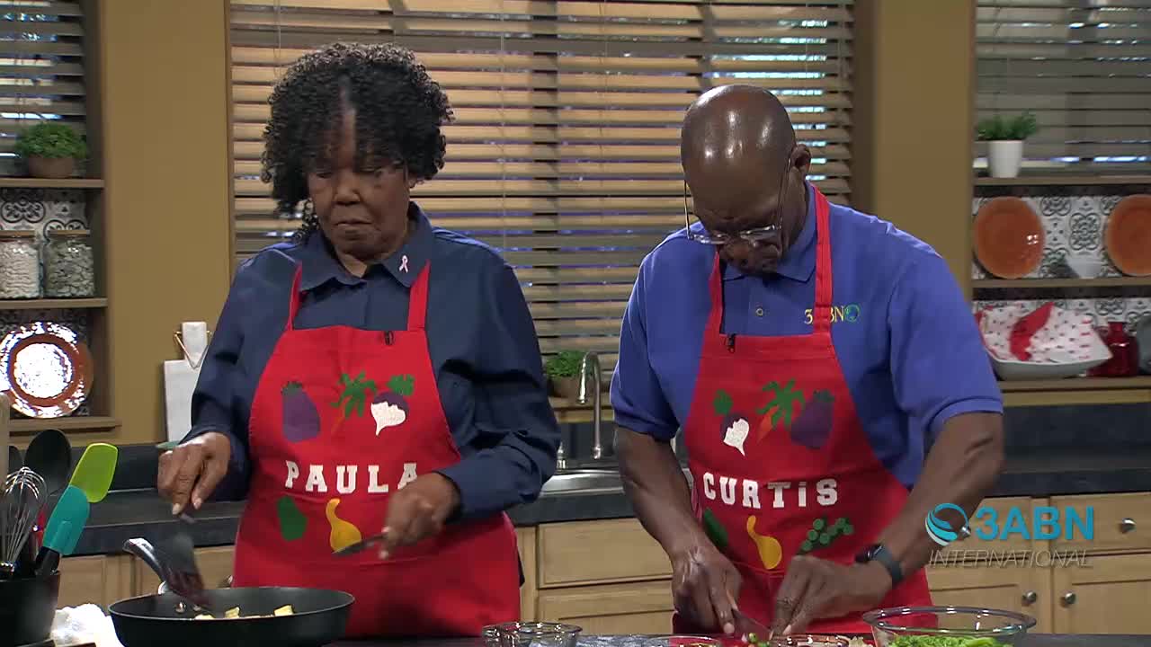 Paula is stirring something in a pan while Curtis chops vegetables. They are both wearing red aprons with their names on them.