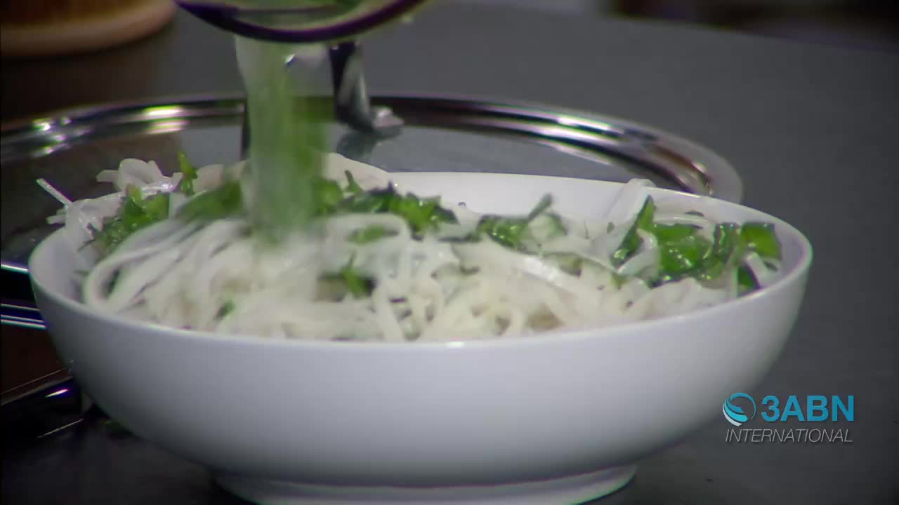 A stream of bright green liquid pours from a metal pitcher into a white bowl filled with white noodles and chopped green herbs. The 3ABN International logo appears in the lower right corner.