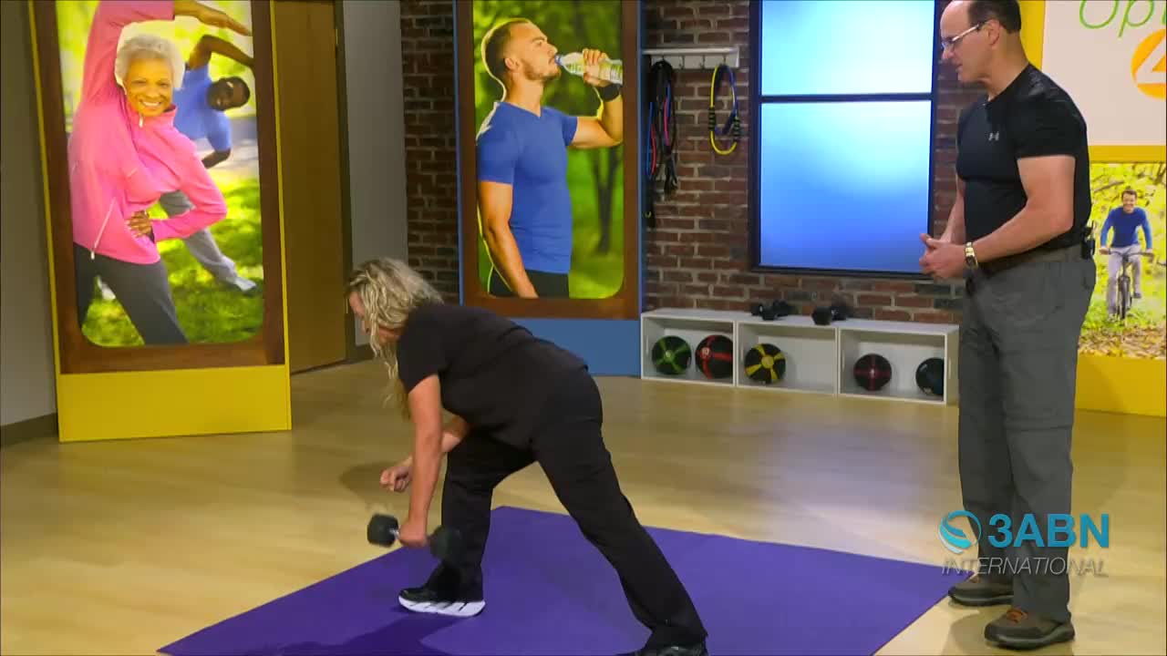 A woman on a purple mat is performing a dumbbell row, her body bent forward. A man stands nearby, observing her workout.