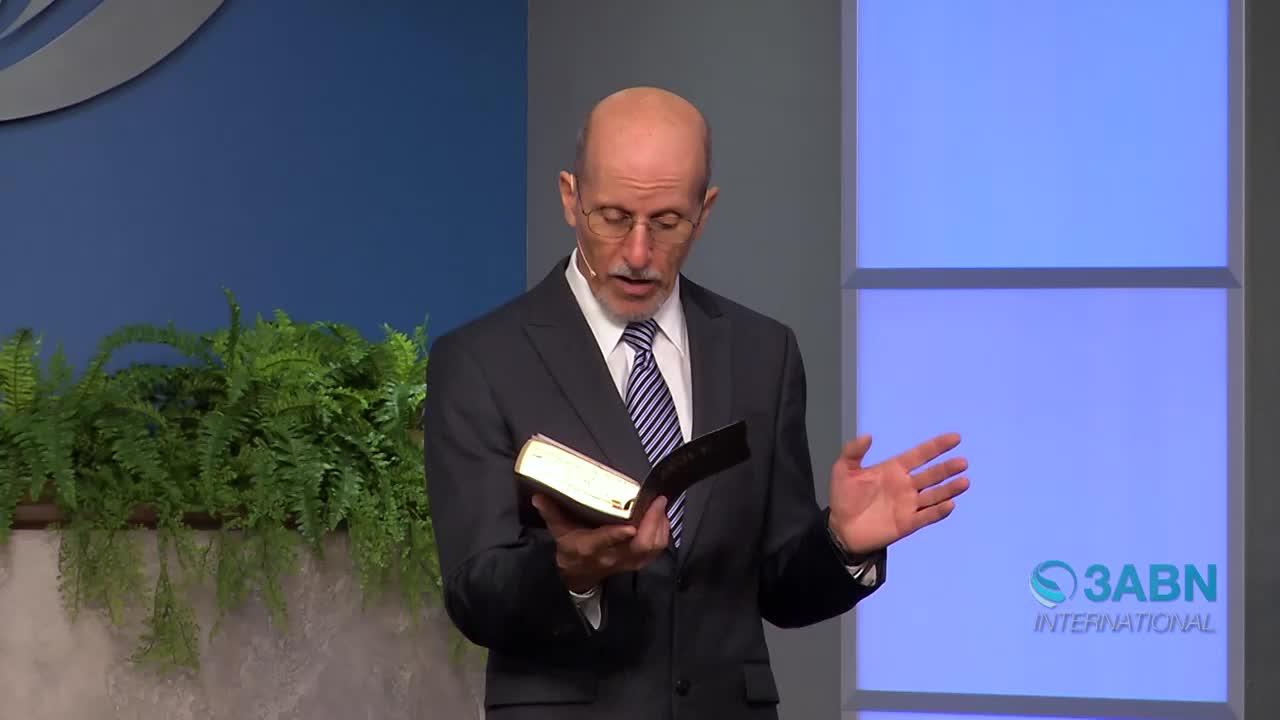 A man in a suit stands on a stage, holding an open Bible. He gestures with his free hand as he speaks, his gaze fixed on the pages. Lush green ferns flank the stage behind him, and the 3ABN International logo is visible on a blue backdrop.