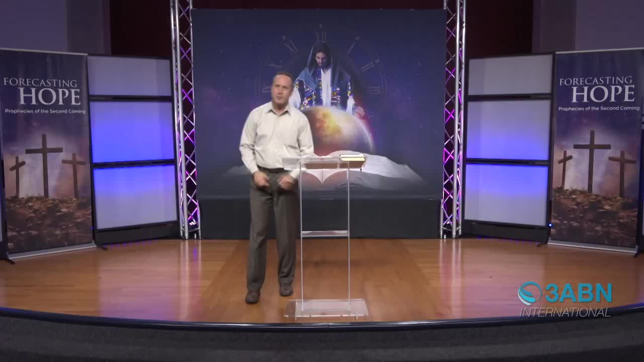 A man stands at a podium on a wooden stage, speaking into a microphone. Behind him, a large screen displays an image of Jesus with a globe. Banners on either side of the stage read "Forecasting Hope: Prophecies of the Second Coming."