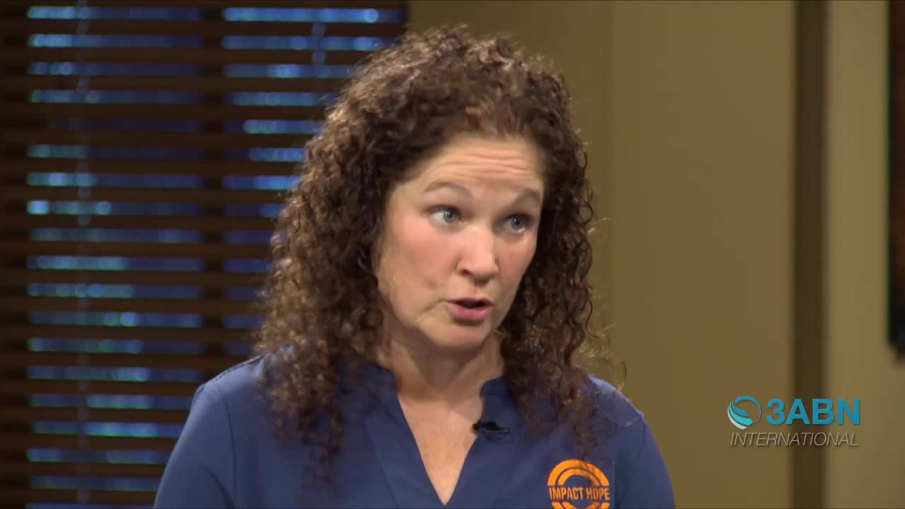 A woman with curly brown hair speaks directly to the camera. She wears a blue shirt with an "Impact Hope" logo.