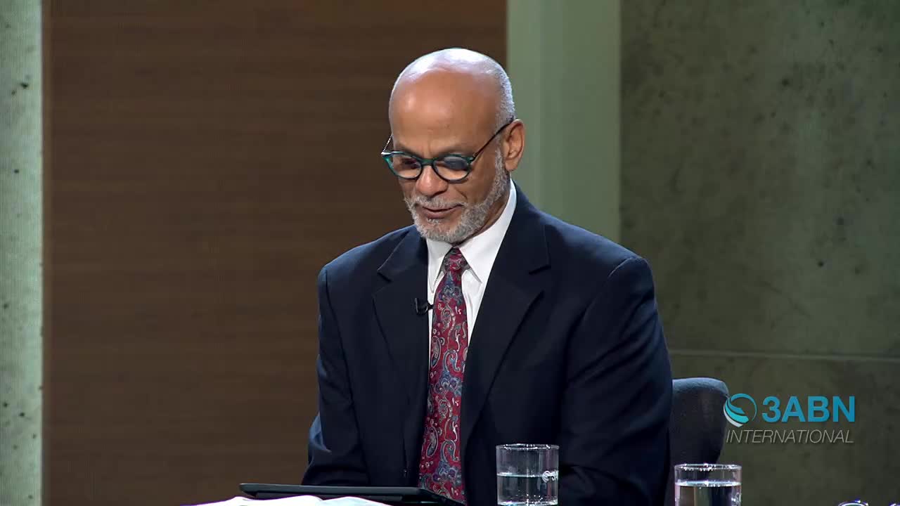 A man in a dark suit and patterned tie looks down, his glasses perched on his nose. A glass of water sits on the table before him, reflecting the studio lights.