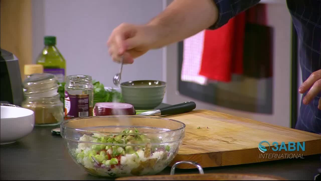 A hand sprinkles white powder from a small spoon into a clear bowl of chopped vegetables. On the counter beside it, a red onion and a jar of spices sit ready for use.