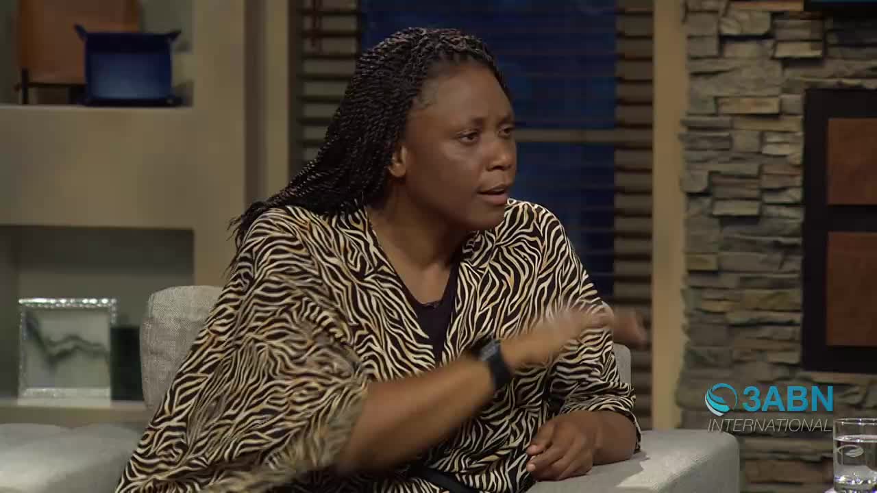 A woman with braided hair gestures with her right hand as she speaks. She wears a black shirt under a patterned blouse, sitting on a couch in what appears to be a studio setting.