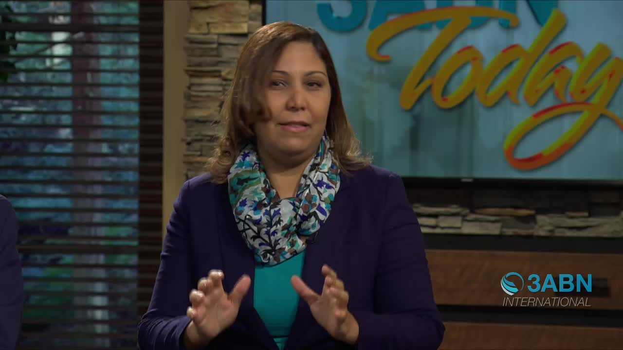 A woman with auburn hair, wearing a dark blue jacket and a patterned scarf, gestures with her hands. Behind her, a screen displays "3ABN Today" in large, stylized yellow lettering.