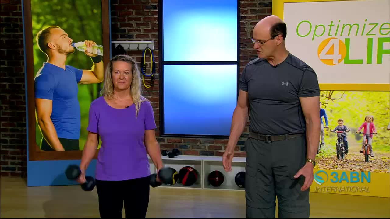 A woman in a purple shirt is doing bicep curls with dumbbells. Beside her, a man in a grey shirt gestures as he speaks, with a backdrop showing the 3ABN International logo and a scene of children biking.