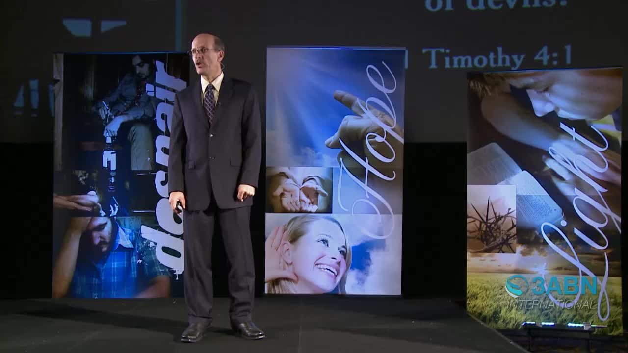 A man in a suit stands on a stage, speaking. Behind him, three large screens display images and text related to 3ABN International.