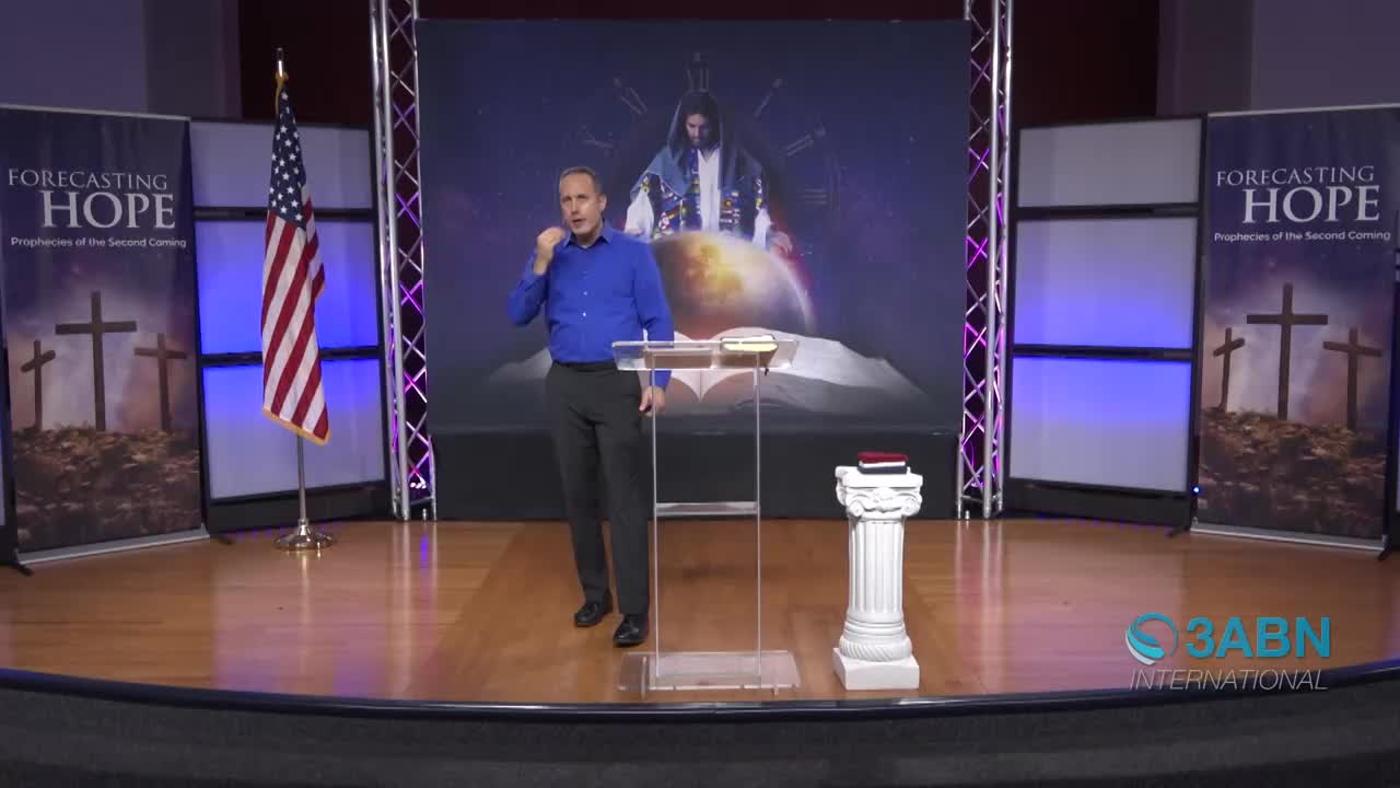 A man in a blue shirt gestures emphatically while speaking from behind a clear podium. Behind him, a large screen displays an image of Jesus with a globe and an open Bible.