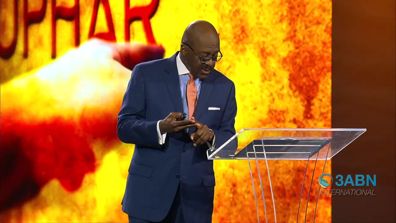 A man in a dark suit stands at a clear podium, gesturing with his hands as he speaks. Behind him, a large screen displays the word "UPHAR" in bold red letters against a fiery yellow and orange background, with the 3ABN International logo visible on the right.