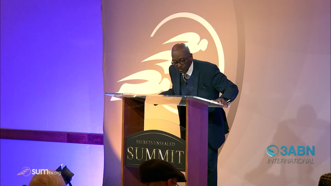 A man in a suit leans over a podium, his hands resting on the clear surface. The backdrop features a large, stylized design and the 3ABN International logo.