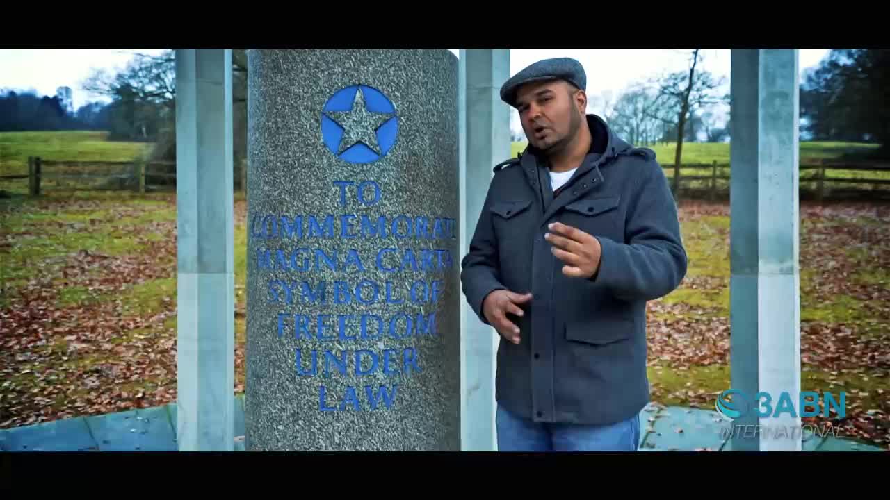 A man in a grey jacket and cap stands beside a stone monument with blue lettering. He gestures with his hands as he speaks, with a grassy field and trees behind him.