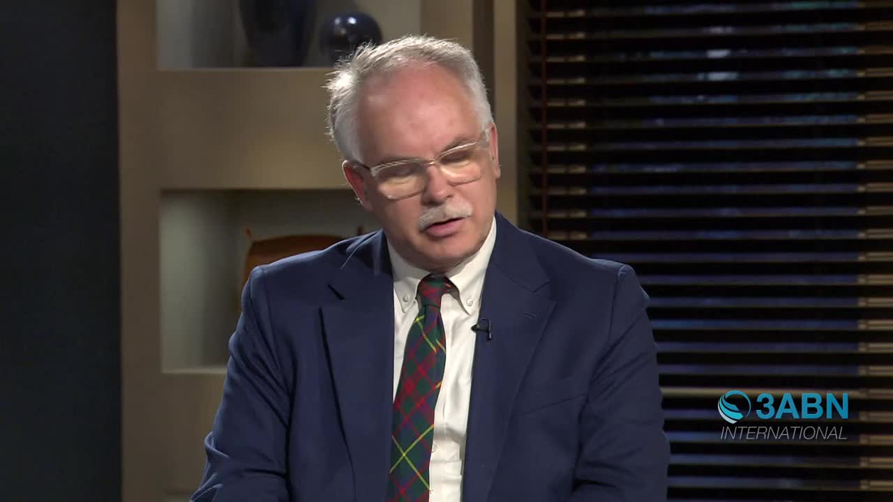 A man in a navy suit and a plaid tie speaks, his glasses reflecting the studio lights. The 3ABN International logo appears in the lower right corner.