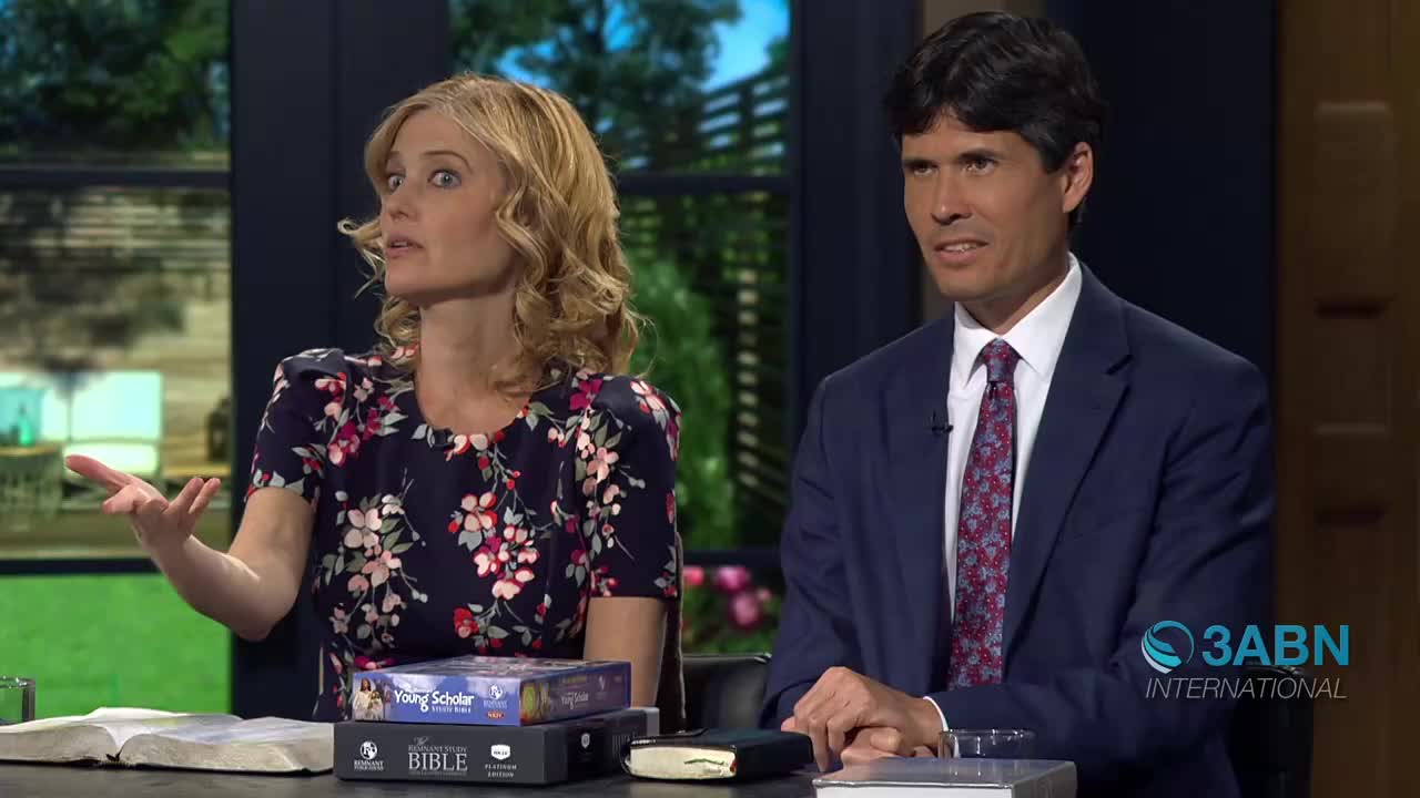 A woman with blonde, curly hair gestures with her left hand while speaking animatedly. Beside her, a man in a navy suit and patterned tie looks slightly off-camera.