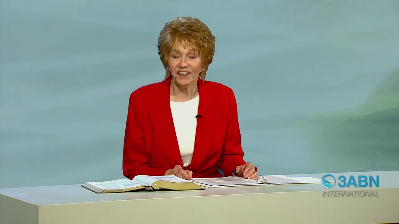 A woman in a red blazer speaks, her hands resting on an open Bible and papers. The 3ABN International logo appears on the right side of the desk.