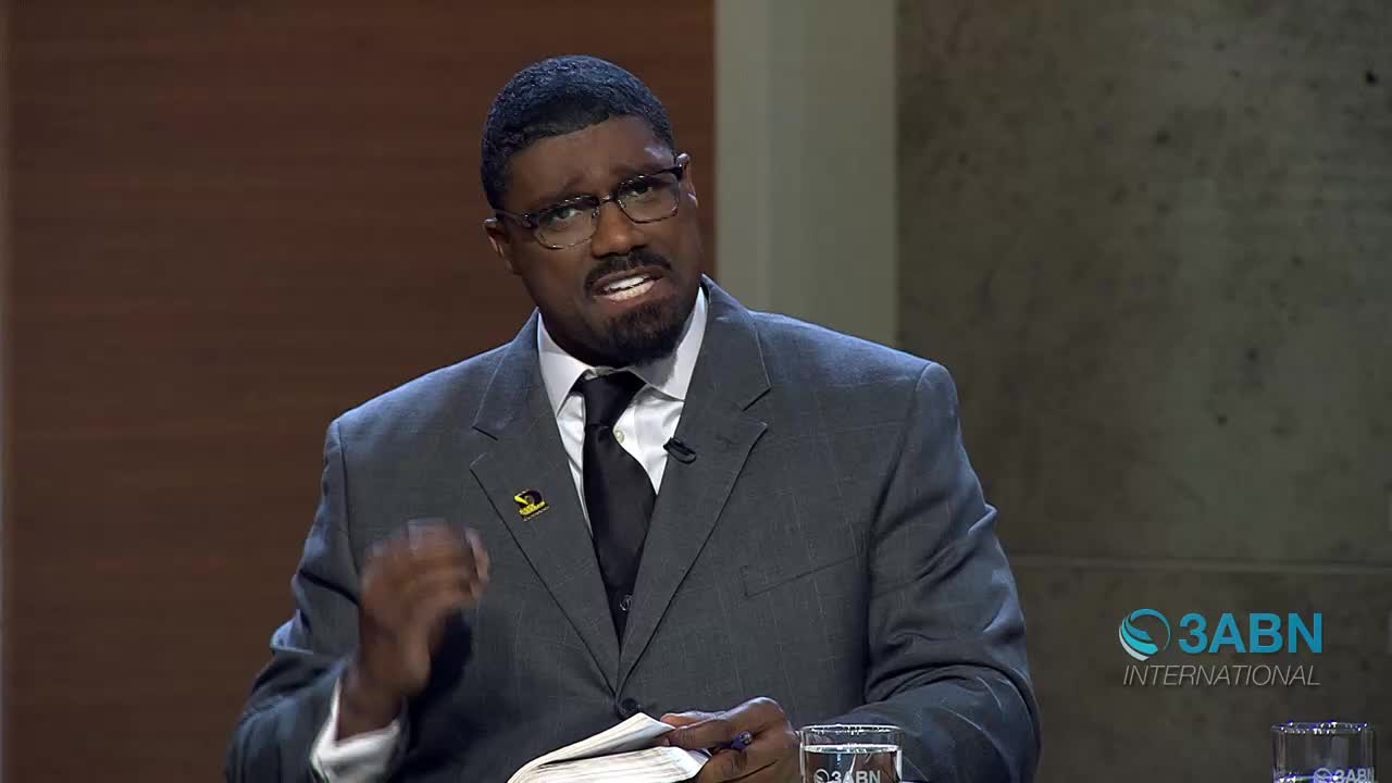 A man in a suit speaks with his left hand raised, gesturing emphatically. He holds a book open in his right hand, and a glass of water sits on the table before him.