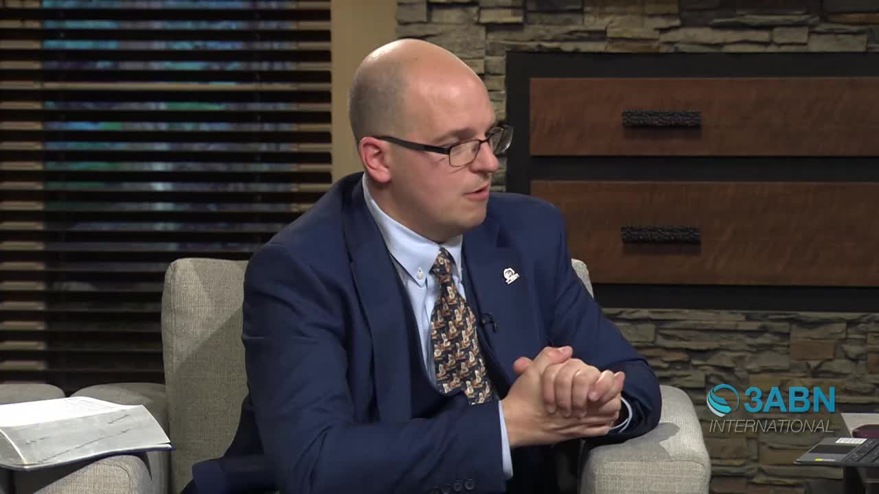A man in a suit and tie speaks, his hands clasped. Beside him, a large book rests on a chair. The 3ABN International logo appears in the corner.