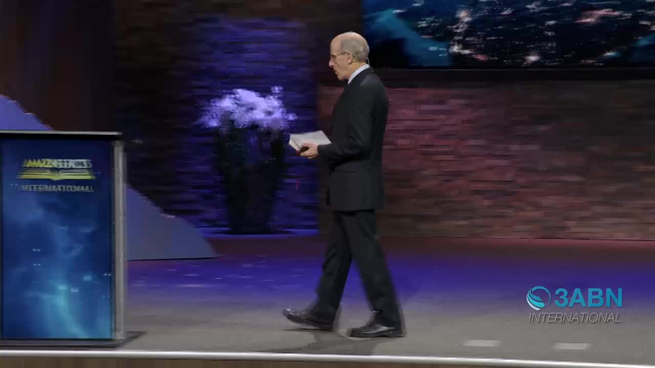 A man in a suit walks across the stage, holding a book. The 3ABN International logo is visible on the right side of the screen.