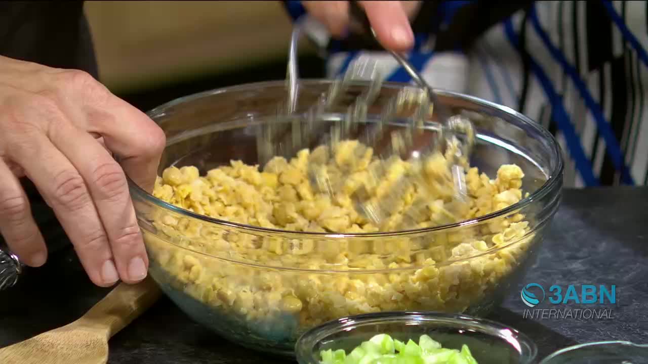 A metal masher is pressing down on a bowl of yellow corn kernels. The corn is being broken down, preparing it for a recipe that might be featured on a 3ABN International broadcast.