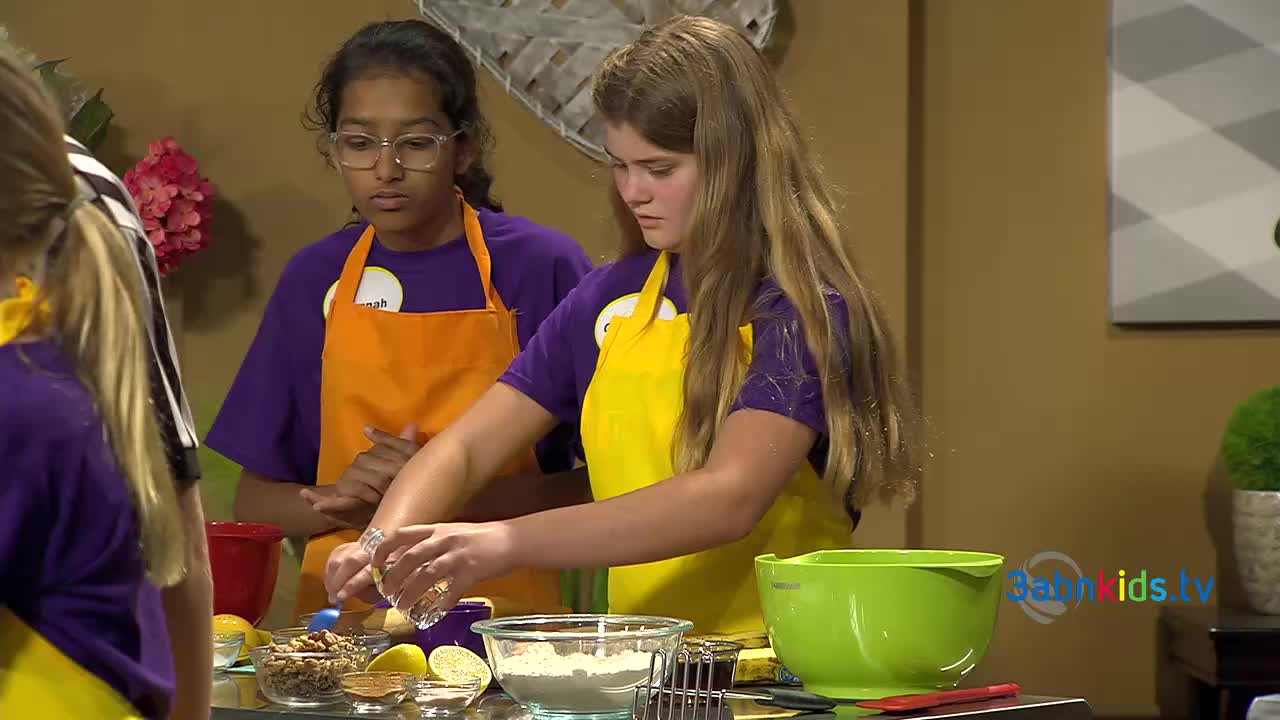 Two girls in aprons and matching shirts are gathered around a counter, working with ingredients. One girl is carefully sprinkling something from a small container into a bowl of flour.
