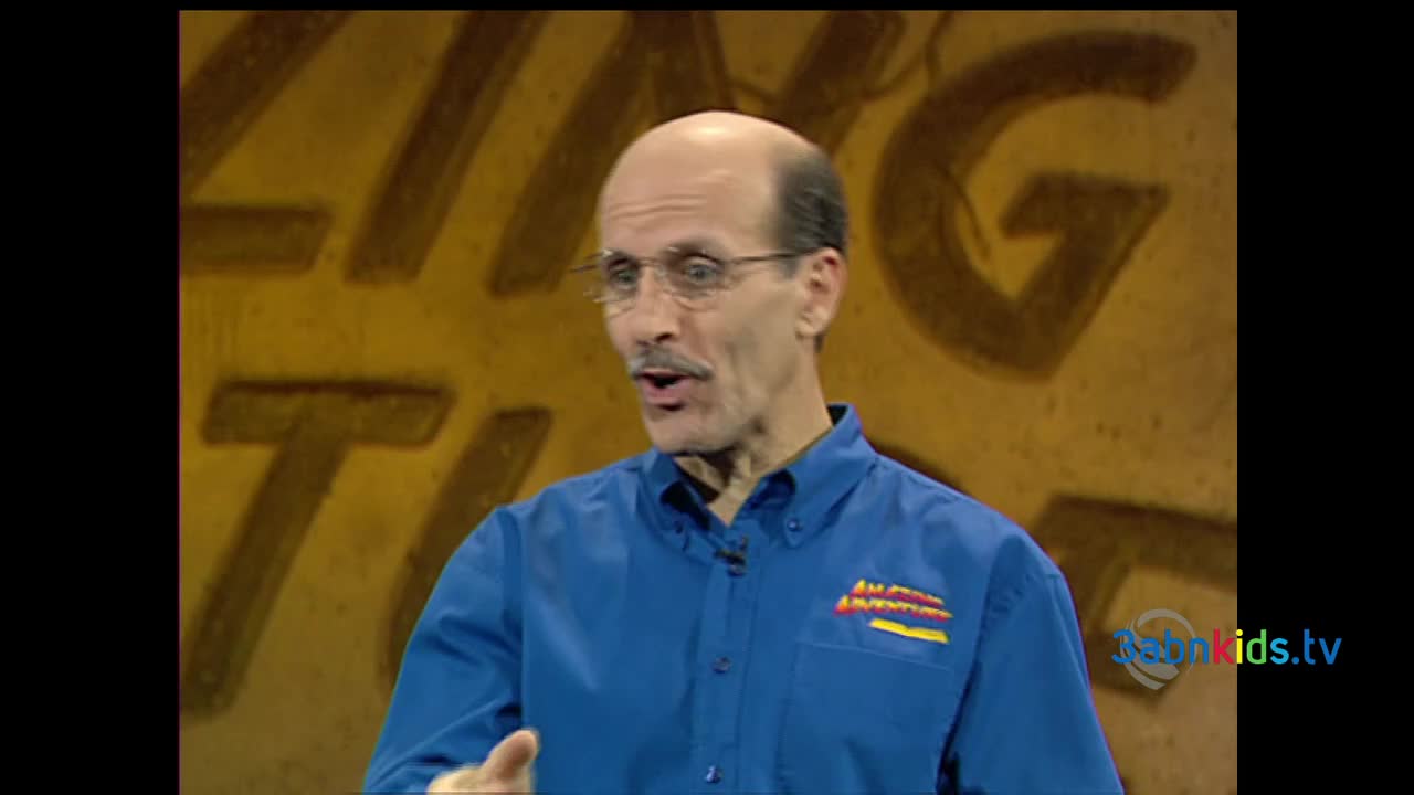 A man in a blue shirt, from what looks like a 3ABN Kids show, is speaking with his hands. Behind him, the backdrop is a golden color with large, stylized letters.
