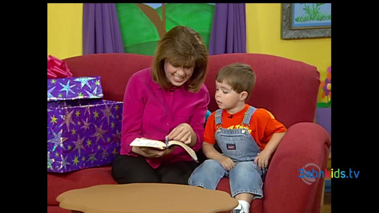 A woman in a pink shirt and a young boy in overalls sit side-by-side on a red couch, looking at a book. The woman smiles as she points to the pages, and the boy watches intently.
